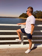 Man wearing Thirsty Threads men’s hybrid boardshorts in navy, standing on a pier by the water.