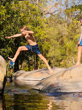 Boy wearing Thirsty Threads Hybrid Boardshorts jumping off a rock, surrounded by trees.