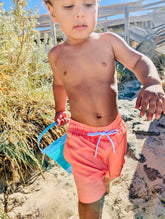Boy in Thirsty Threads Burnt Horizon Lined Hybrid Boardshorts standing on a sandy beach with grass and holding a blue bucket