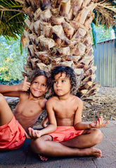 Two brothers sitting under a palm tree, giving thumbs up wearing Thirsty Threads Hybrid Booardshorts in Burnt Horizon.