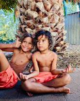 Two brothers sitting under a palm tree, giving thumbs up wearing Thirsty Threads Hybrid Booardshorts in Burnt Horizon.