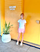 Girl wearing Thirsty Threads pink striped hybrid girl's boardshorts and white tee, standing by yellow wall at Sunshine Social