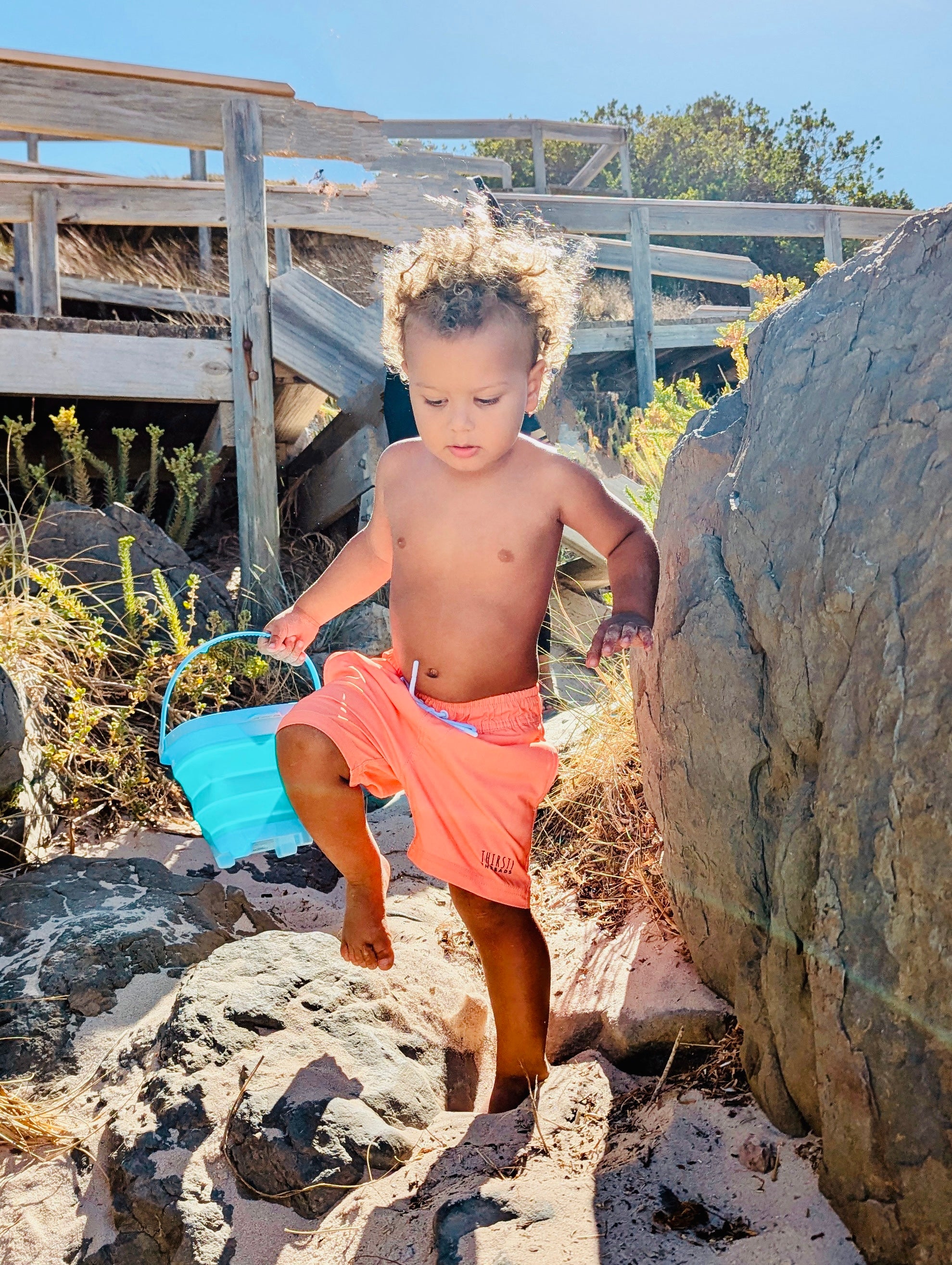 Child playing on rocks at the beach with a blue bucket wearing Thirsty Threads Hybrid Boardshorts in Burnt Horizon