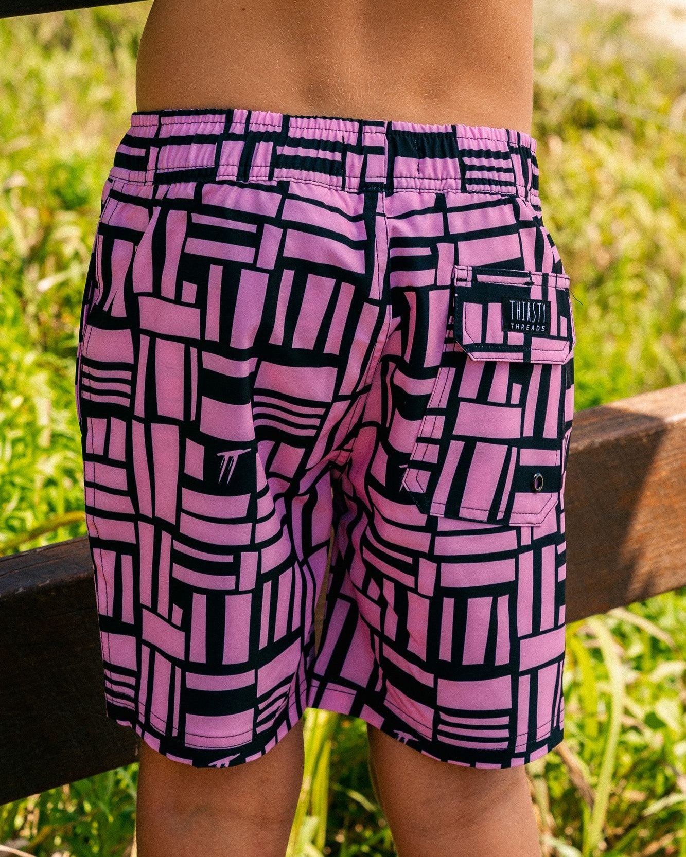Child wearing Thirsty Threads boys pink geo anti-chafe hybrid boardshorts at the beach entrance - back view